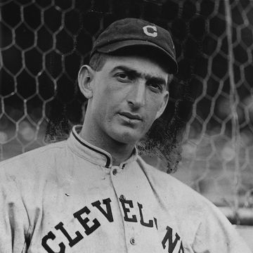 a man stands in a cleveland baseball uniform and hat and stares at the camera, a chain link fence is behind him