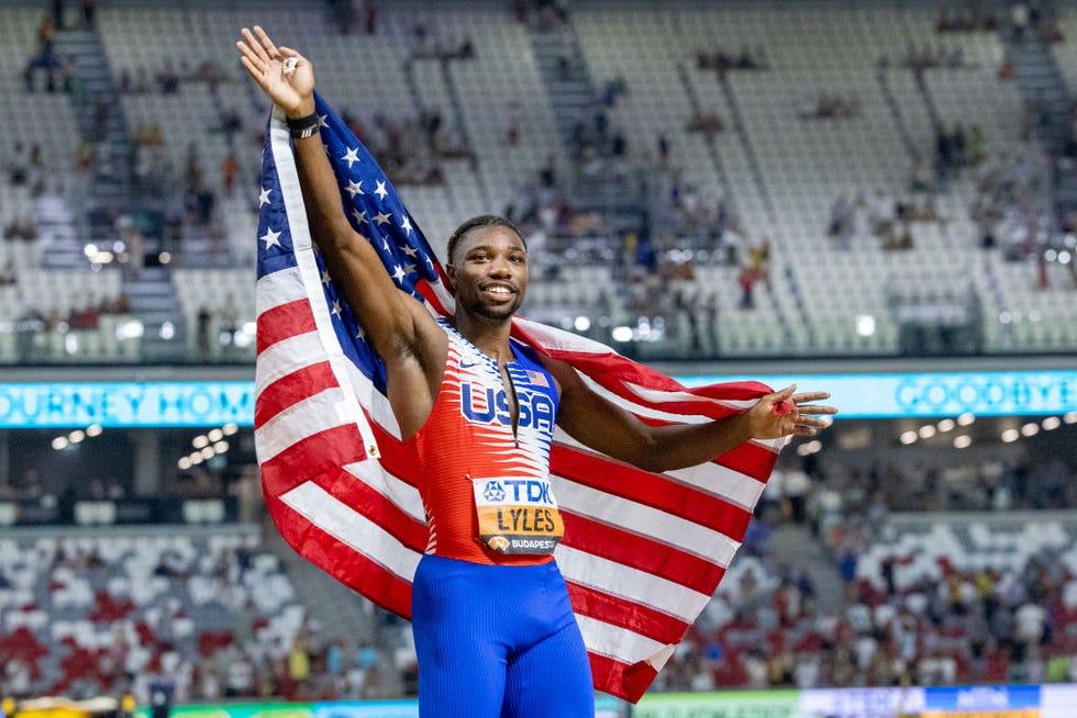 noah lyles smiles and waves as he holds an american flag across his back, he wears a red white and blue uniform and a bib with his name on it, behind him is a stadium partially filled with fans