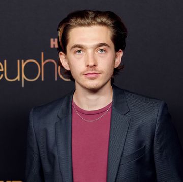 austin abrams, a young man stands looking at the camera, brown hair slicked back, wearing red tshirt and black jacket