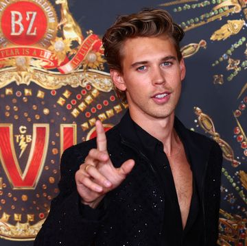 austin butler wearing a black shirt, holding a finger in the air, and standing in front of a logo with the word elvis on it