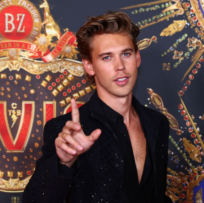 austin butler wearing a black shirt, holding a finger in the air, and standing in front of a logo with the word elvis on it
