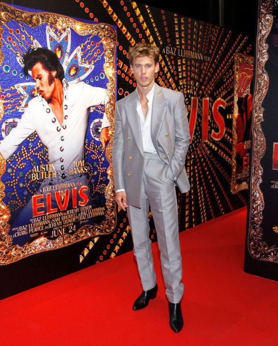 austin butler wearing a silver suit and standing next to a film poster showing him in costume as elvis presley