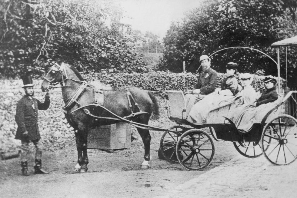 charles dickens sits in the front of a carriage next to his wife catherine hogarth dickens, two other girls are also seated in the carriage, a man wearing a tall top hat stands next to the horse attached to the carriage