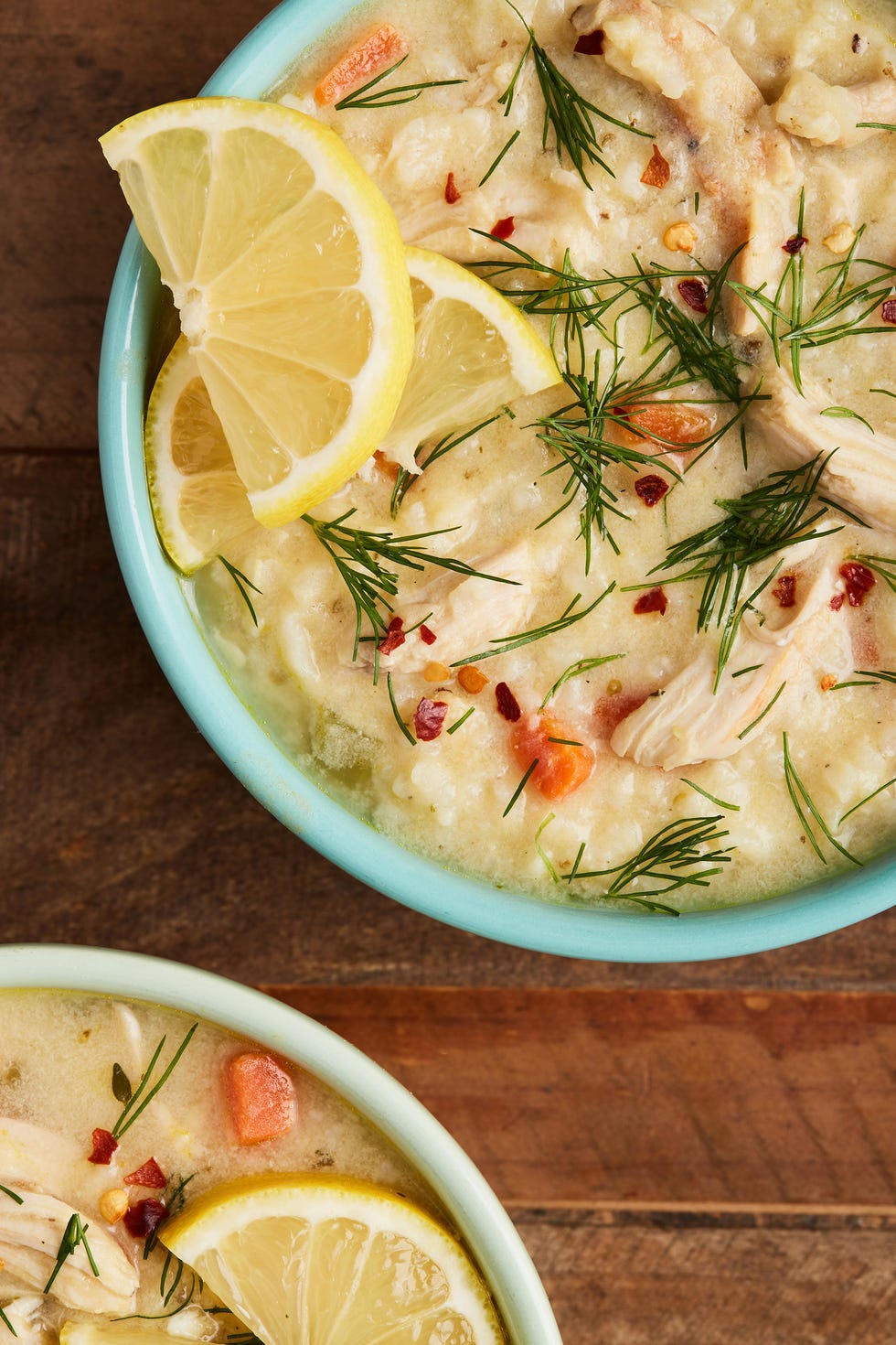 two teal bowls of avgolomeno soup garnished with red pepper flakes, lemon wedges, and dill