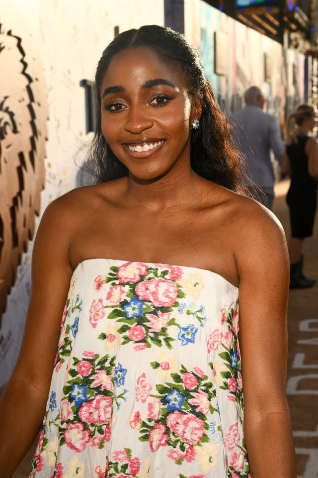 ayo edebiri stands outside and smiles at the camera, she wears a strapless floral print dress