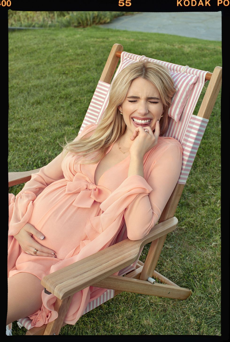 emma roberts, pregnant, wearing peach dress, sitting outside in a striped pink and white lawn chair on the grass