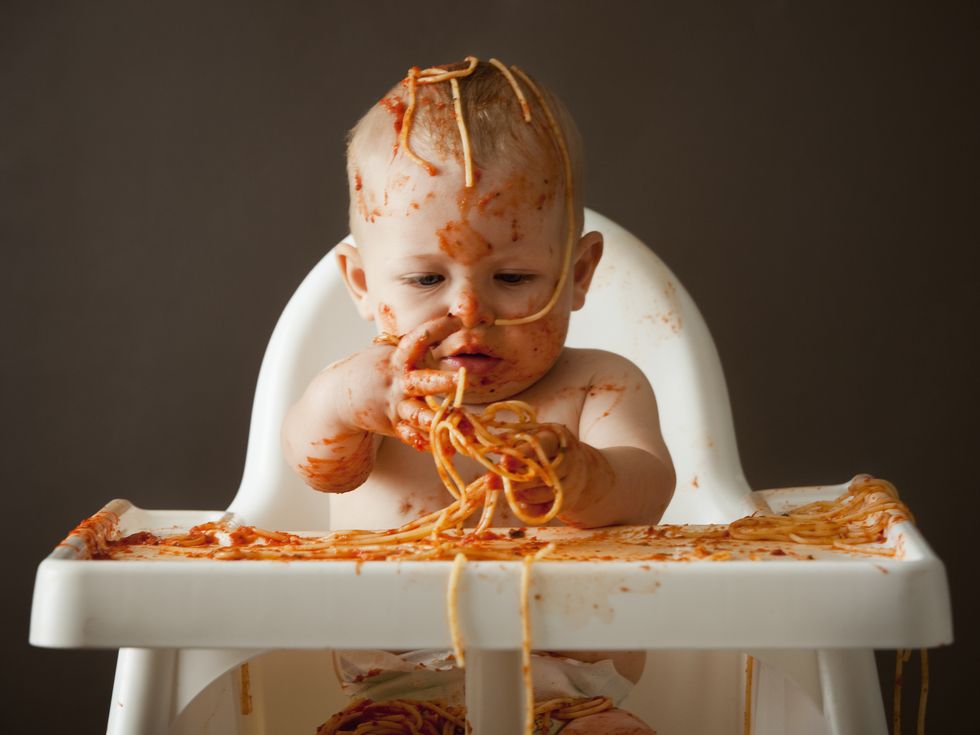 bebé comiendo espaguetis sentado en la trona y cubierto de tomate