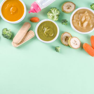 infant baby food bowls with vegetable fruit puree, green, orange, yellow colors   broccoli, carrots, banana, apple with baby accessories and toys green background flat lay top view copy space