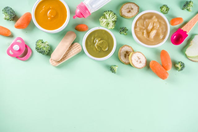 infant baby food bowls with vegetable fruit puree, green, orange, yellow colors   broccoli, carrots, banana, apple with baby accessories and toys green background flat lay top view copy space