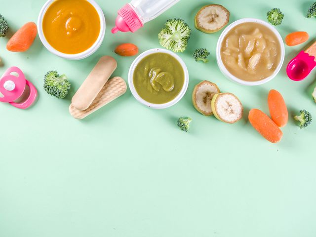 infant baby food bowls with vegetable fruit puree, green, orange, yellow colors   broccoli, carrots, banana, apple with baby accessories and toys green background flat lay top view copy space