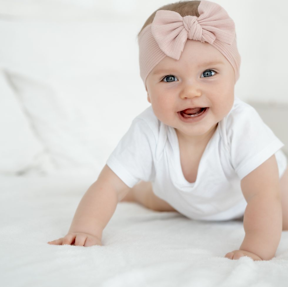 multicultural baby names, a baby girl is smiling at home on a white bed with a bow on her head, a happy healthy child of six months is laughing