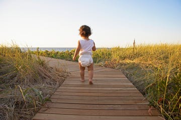 baby girl 12 15 months walking on boardwalk amongst grass, rear view