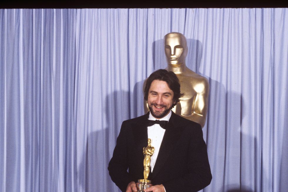 robert deniro, wearing a black tuxedo, smiling and holding an oscar statuette and standing in front of a purple curtain and a large oscar statue