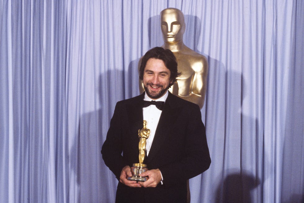 robert deniro, wearing a black tuxedo, smiling and holding an oscar statuette and standing in front of a purple curtain and a large oscar statue