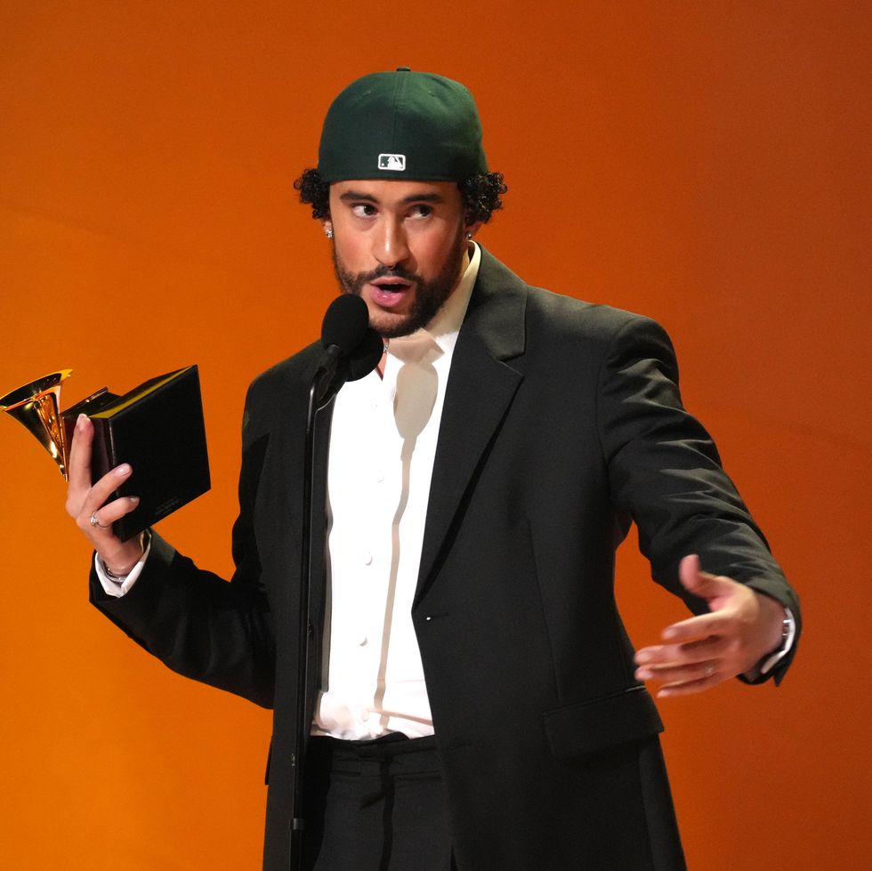 a man in a black suit and white collard shirt with a backward ball cap speaks into a microphone on a stand while accepting a grammy award