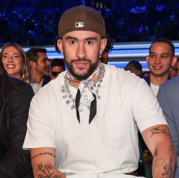 bad bunny looks at the camera while sitting down next to people, he wears a white t shirt and jeans with jewelry and a backward baseball cap