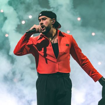 a man in a black outfit and backward ball cap with an orange top stands on a smoky stage and sings into a microphone he holds with one hand