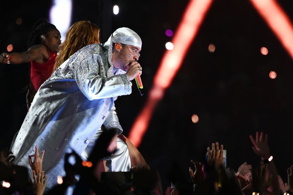 bad bunny, wearing a white and silver sparkling coat and durag, sings into a microphone while kneeling forward on a stage in front of fans raising their hands toward him