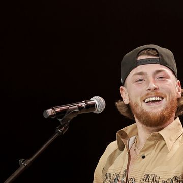 a man in a backward ball cap smiles at the camera while sitting behind a microphone on a stand, he wears an orange button up shirt with a western design