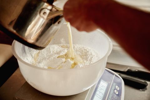 A baker pouring liquid from a copper jug into a bowl with flour and ingredients for dough.