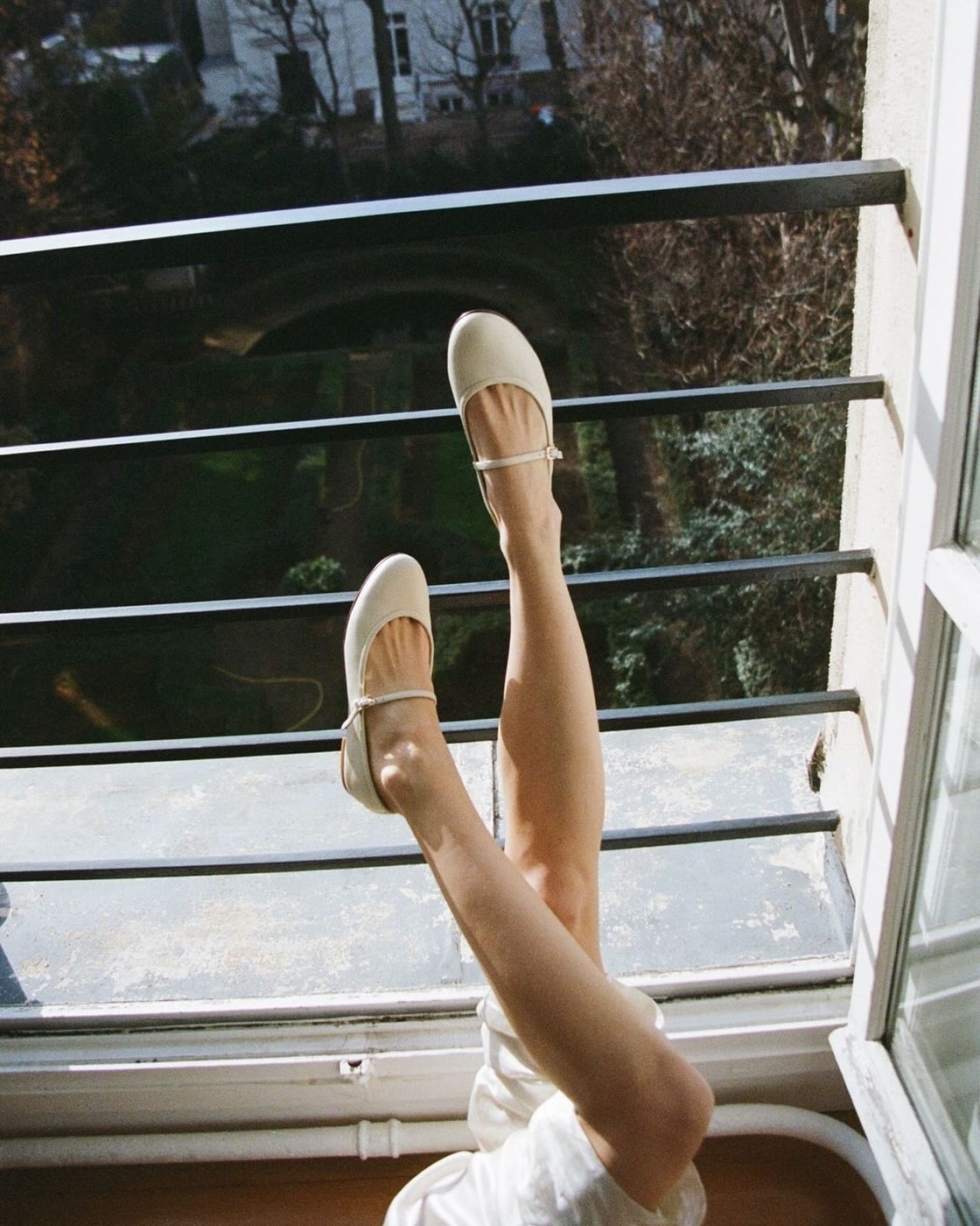a woman wearing ecru mary jane ballet flats by margaux with her legs kicked up on a balcony in a roundup of the best ballet flats for women 2024