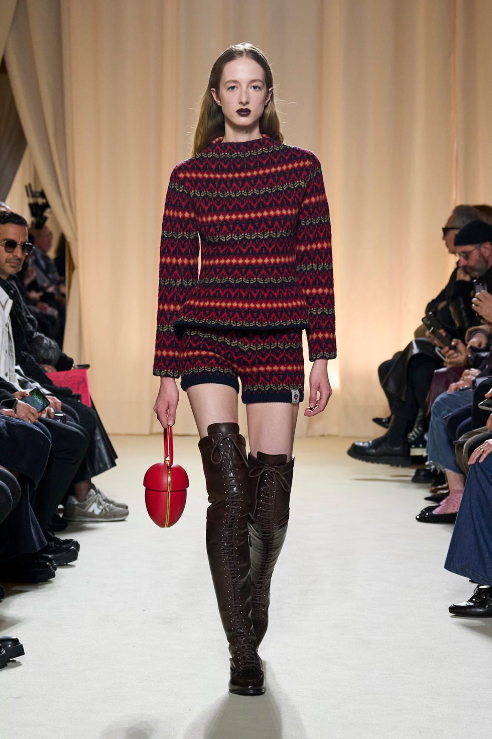 Fashion model wearing a patterned sweater and shorts on a runway holding a red handbag