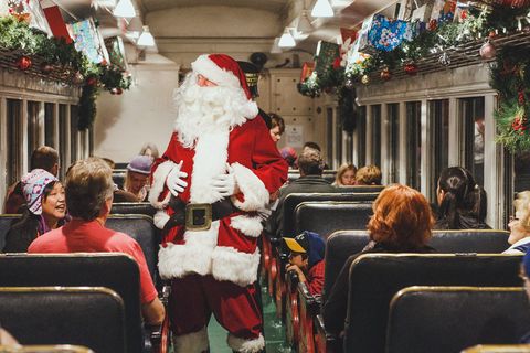 santa is standing in the aisle laughing onboard a vintage train with passengers around him there are swags of christmas greenery, christmas ornaments and presents draped from the ceiling