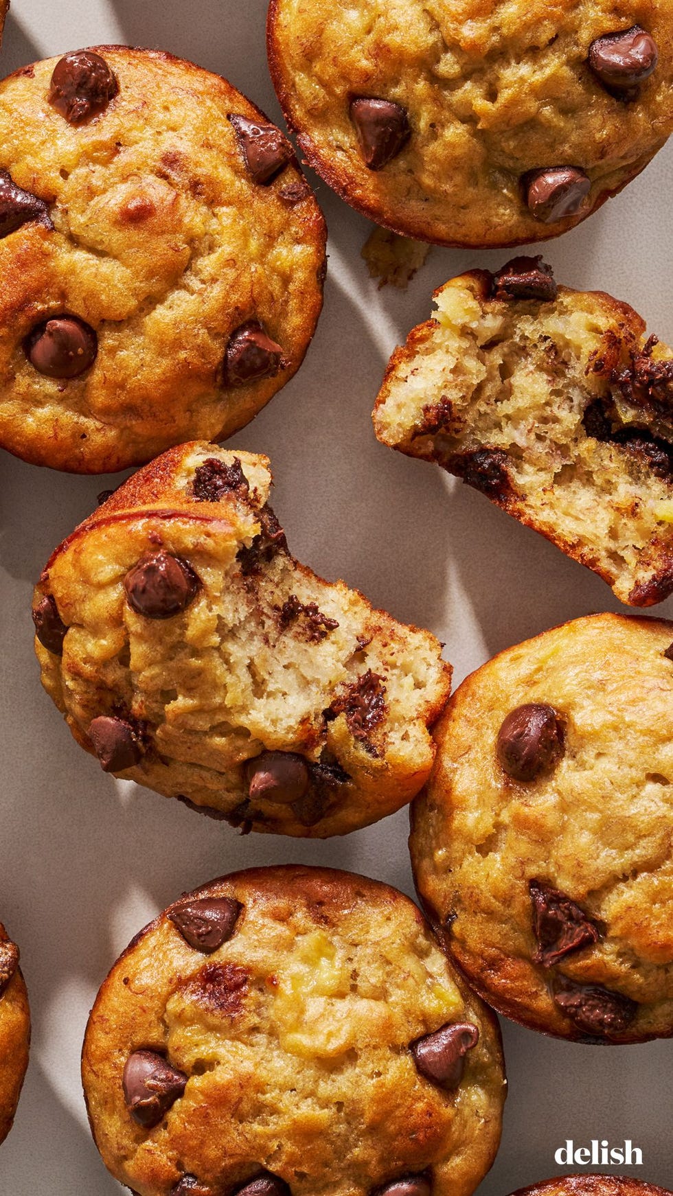four whole banana chocolate chip muffins on a white surface with a fifth muffin torn in half sitting in the center