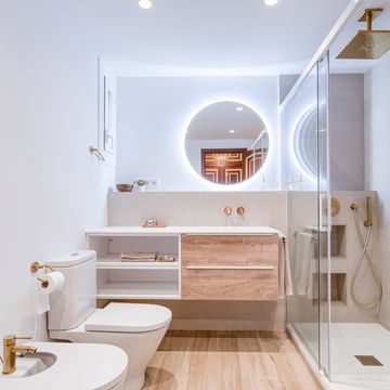 baño con ducha, mueble en blanco y madera volado, espejo redondo y griferías y detalles dorados