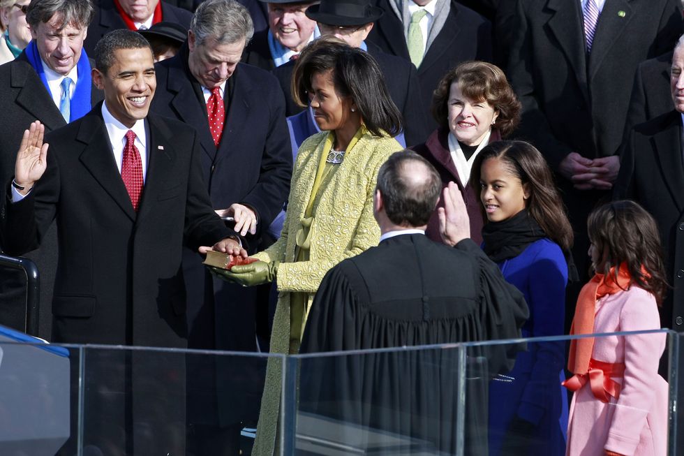 barack obama holds up his right hand and smiles at john roberts while his left hand rests on a bible held by michelle obama, in the crowd around them are malia obama, sasha obama, diane feinstein and others