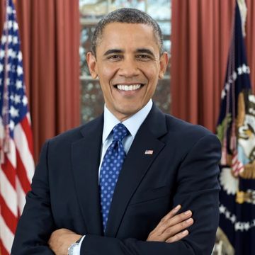 barack obama smiles at the camera with his arms crossed, he is wearing a dark navy suit coat, white collared shirt, blue tied with white polka dots, and an american flag pin on his lap, behind him are sienna curtains, an american flag, and a flag with the presidential seal