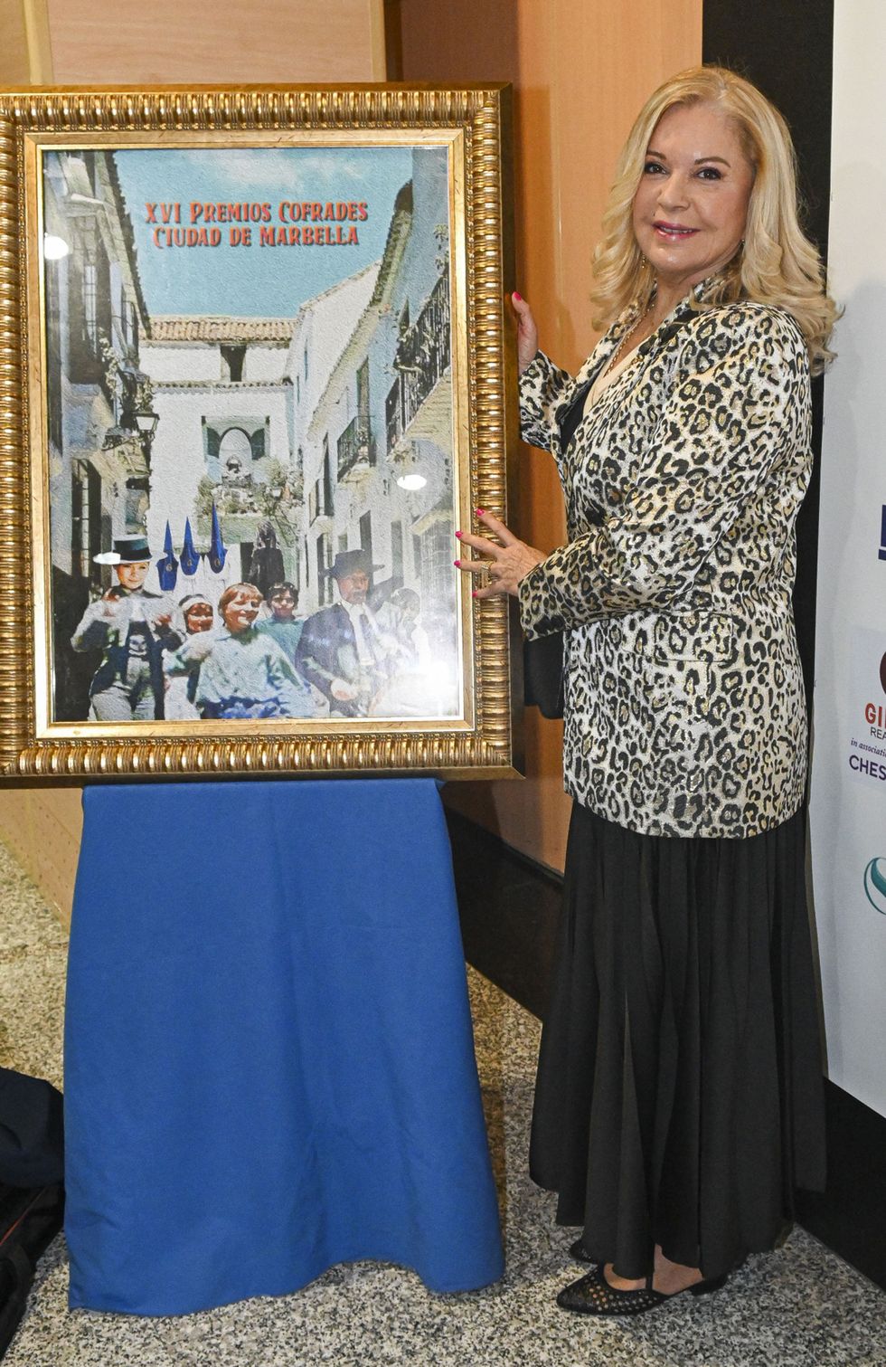la actriz con americana print animal y falda negra posa con el cartel de los premios cofrades ciudad de marbella