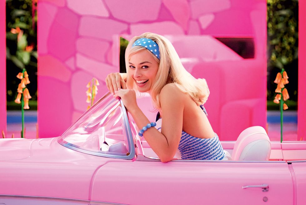 margot robbie, as barbie, sits in a pink car and smiles in a first look photo from the barbie movie