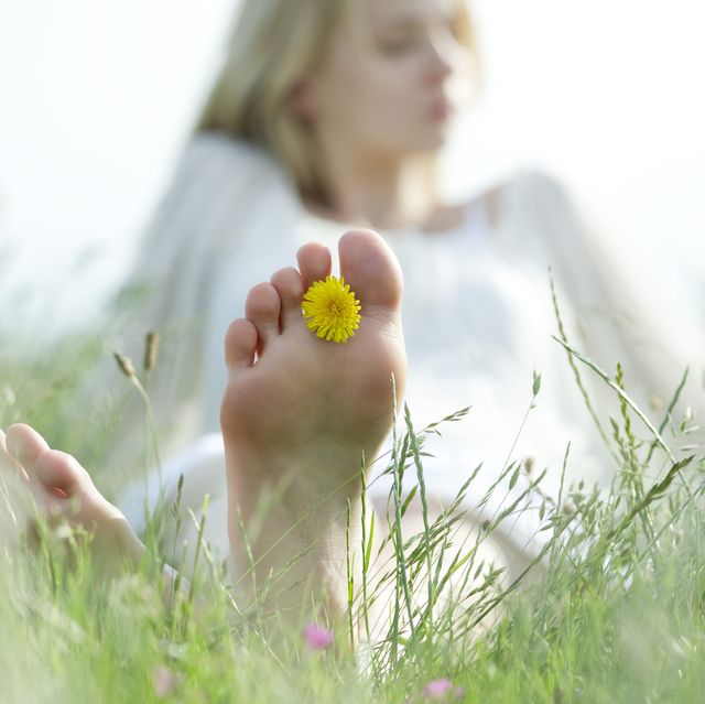 barefoot young woman sitting in grass with dandelion flower between toes, cropped