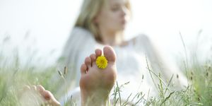 barefoot young woman sitting in grass with dandelion flower between toes, cropped