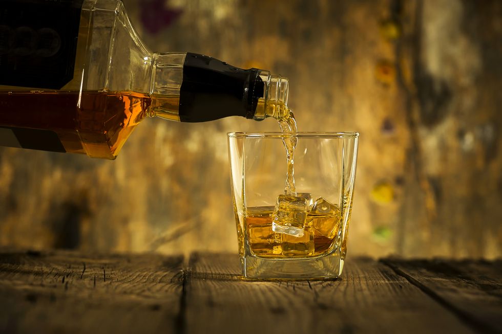 Barman pouring whiskey in front of whiskey glass and bottles on black table with whiskey and ice on a glass table