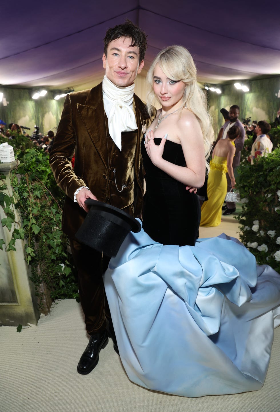 barry keoghan and sabrina carpenter pose for a photo while standing together and hugging, he wears a velvet brown three piece suit with a white high collared shirt and holds a black top hat in one hand, she wears a strapless black gown with a baby blue ruffled bottom