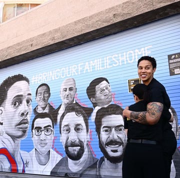 brittney griner hugs someone on her left and looks over her shoulder, she is smiling and both people wear all black outfits, they stand next to a painted mural of 15 people and the slogan bring our families home