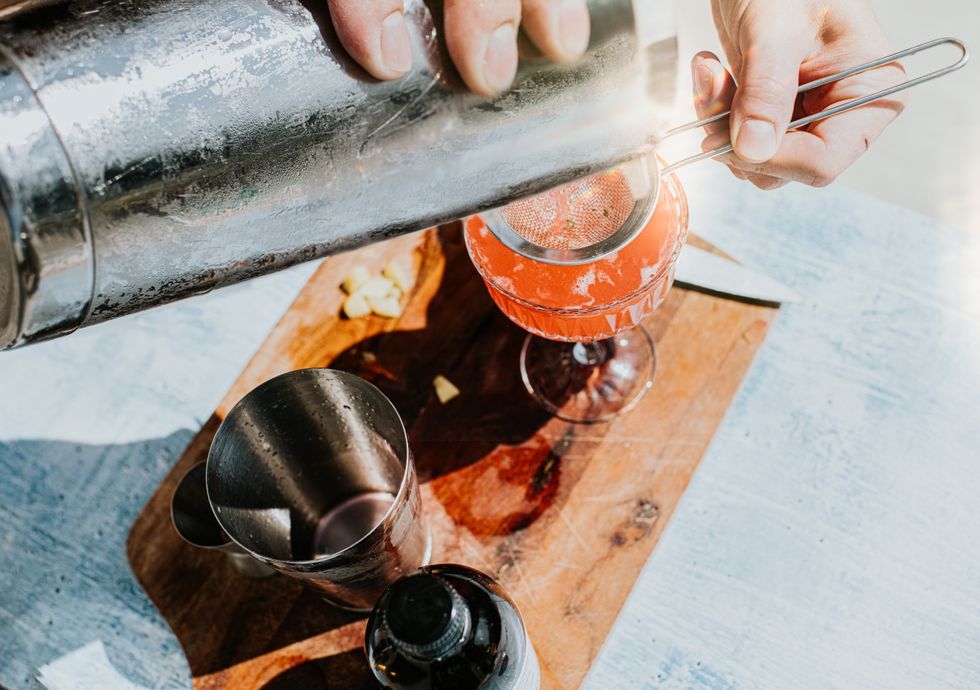 close up of a professional bartender making a refreshing fruit cocktail  mocktail, using a cocktail shaker and strainer he pours the mix through a steel strainer into a glass on a wooden chopping board drink making utensils surround the glass