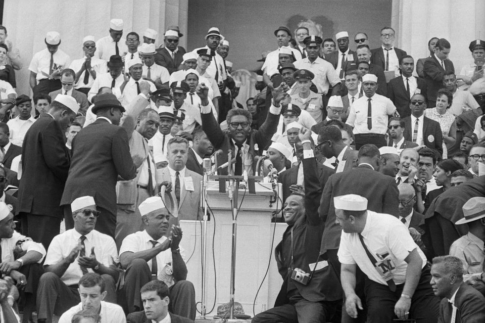 bayard rustin speaks at a podium with his arms raised in the air, people surround him on the steps behind