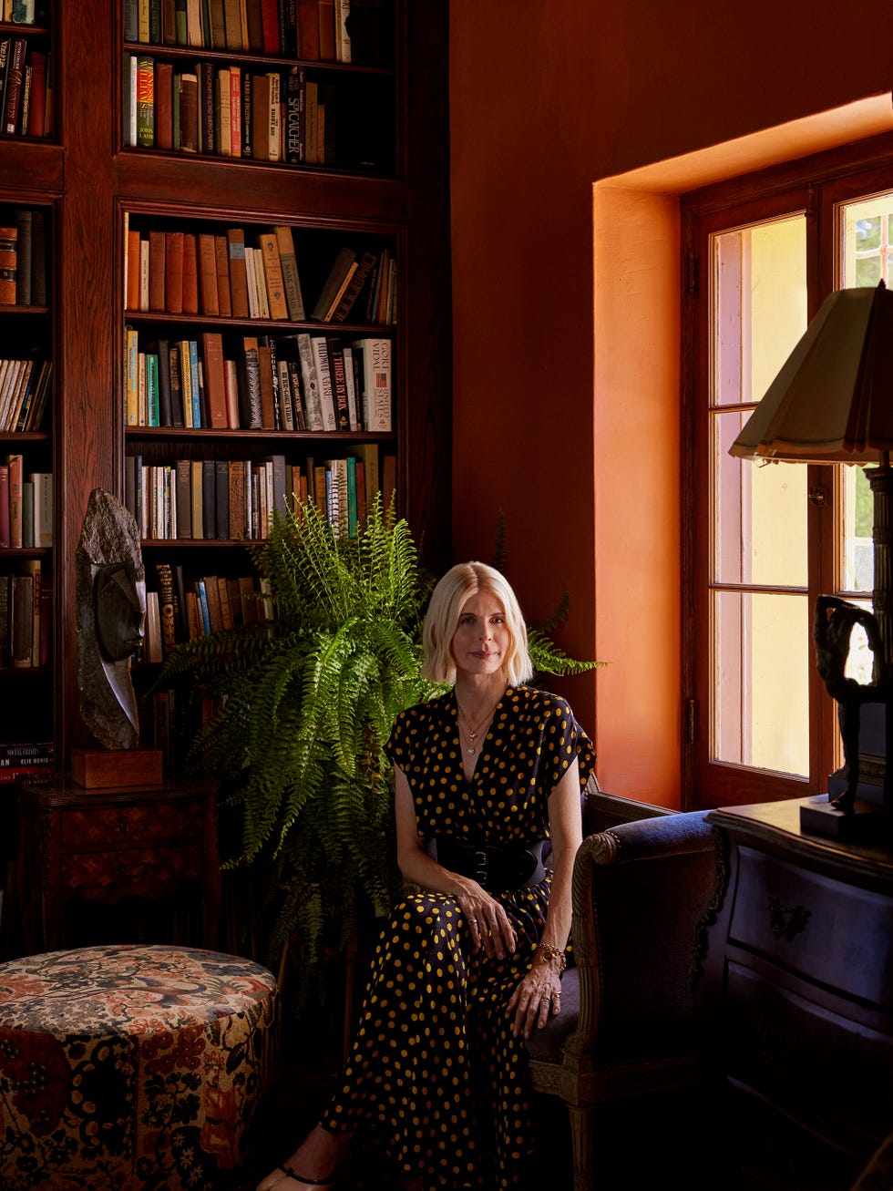 Woman seated in a reading nook with bookshelves and plants.