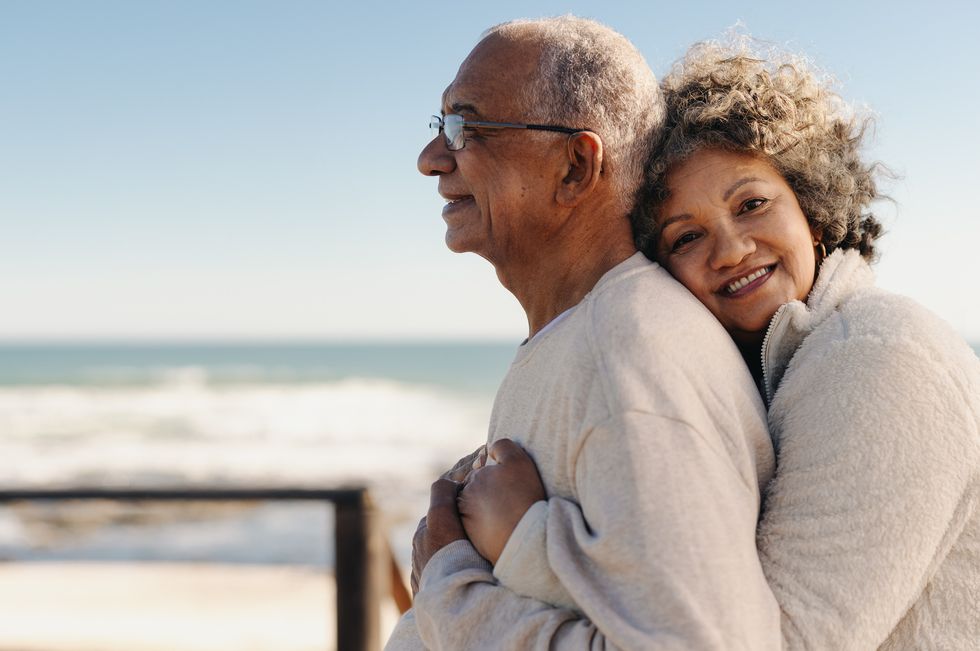 romantic senior woman smiling at the camera while embracing her husband by the ocean affectionate elderly couple enjoying spending some quality time together after retirement