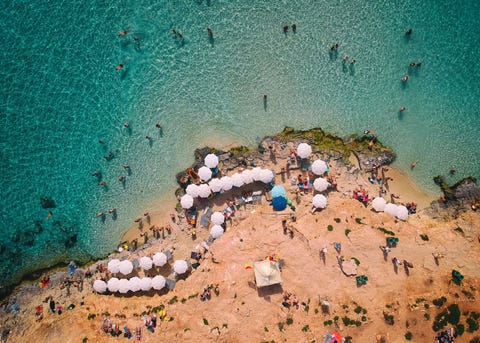Zwemmers waden door het water van een blauwe lagune die tussen de Maltese eilandjes Comino en Cominotto ligt Het 26 vierkante kilometer kleine Comino was ooit een toevluchtsoord voor slachtoffers van de pest en ridders van de Maltezer Orde maar nu is het serene eilandje van kalksteen een plek van ongerepte strandjes vogelkolonies natuurreservaten en bijenkorven