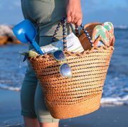 person holding straw beach tote by ocean with shovel flip flops and sunglasses