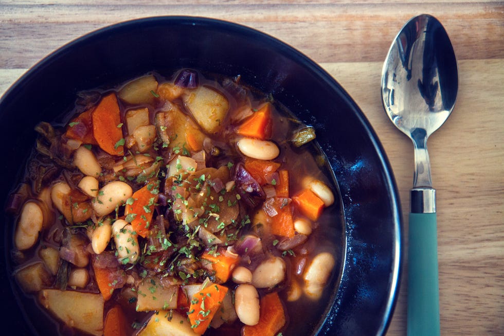 White beans soup with onion, carrot, potatoes and various vegetables in a black plate with a bit of parsley on top and spoon on the side.