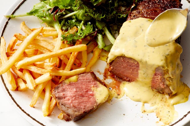 bearnaise sauce being poured over a steak on a plate with salad and fries