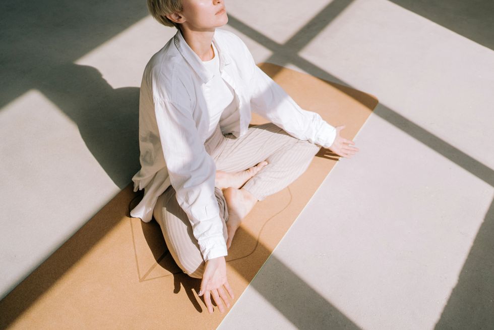 beautiful authentic woman with short blond hair is meditating sitting in lotus position on yoga mat in front of a window she is wearing a light colored casual clothing concept of relaxation exercises
