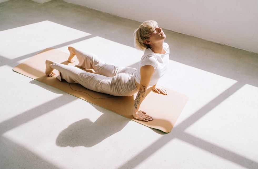 beautiful authentic woman with tattoos and short blond hair is doing yoga in upward facing dog position on yoga mat in front of a window she is wearing a light colored casual clothing concept of relaxation exercises