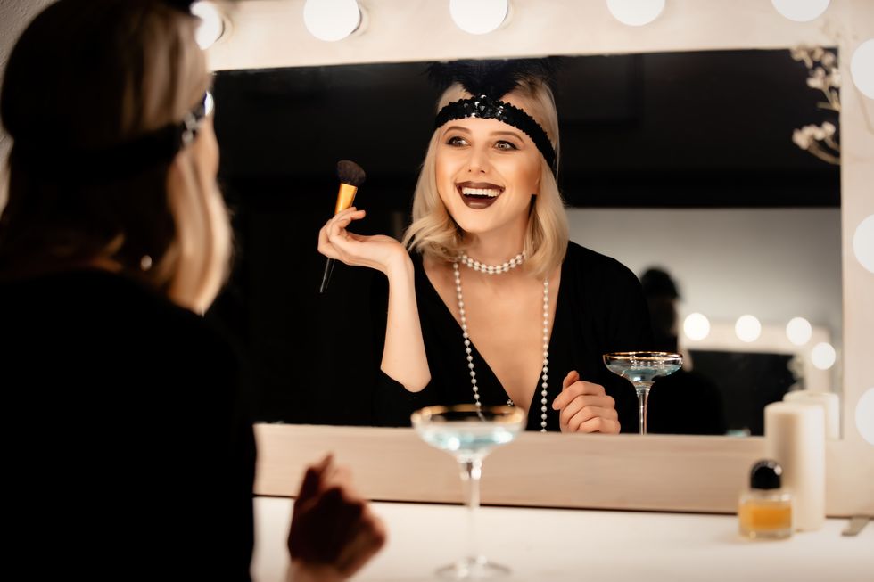 beautiful blonde woman in twenties years clothes applying makeup near a mirror with bulbs
