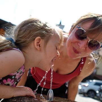 beautiful mid adult mother is drinking water with her daughter at outdoor water fountain in summer